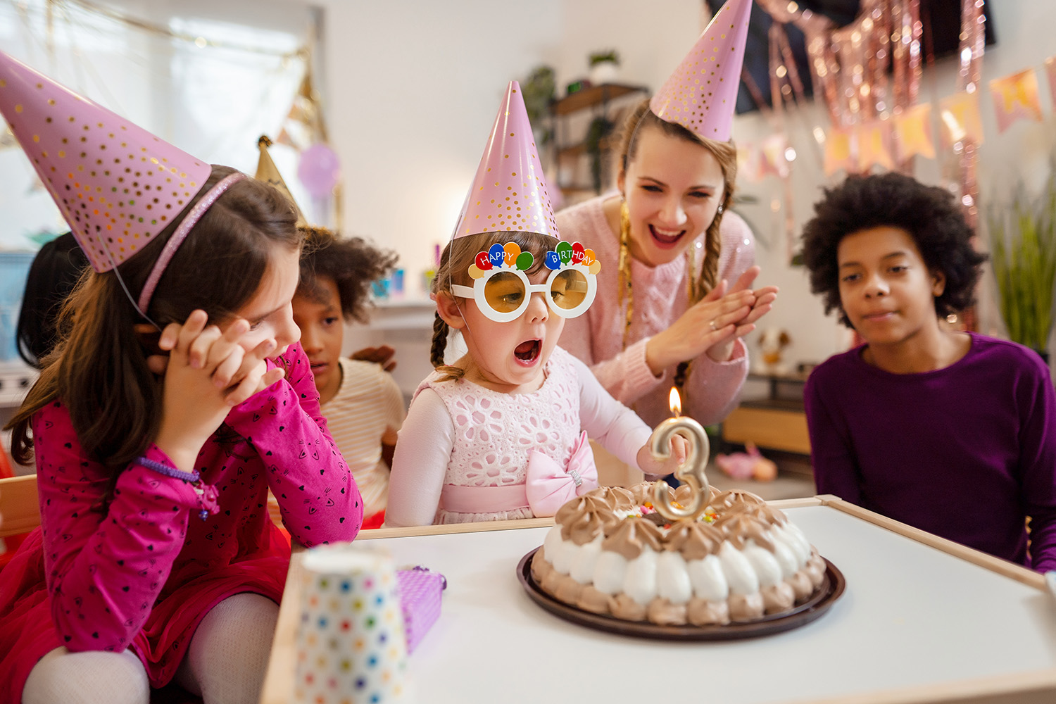 Fyra barn i färgglada festhattar samlas runt en födelsedagstårta med ett ljus format som siffran tre