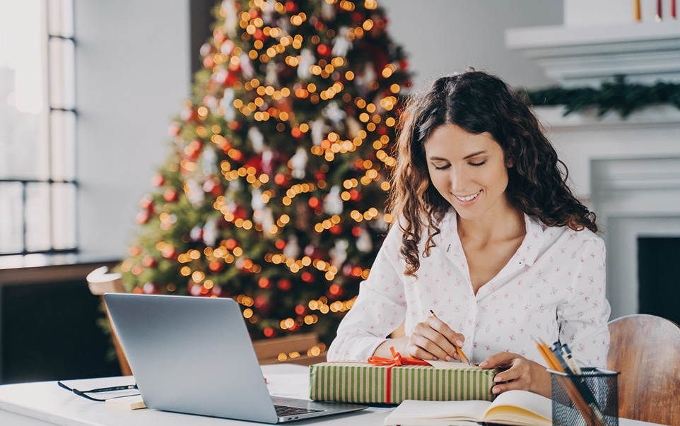 En kvinna som skriver på ett presentkort framför sin laptop, med en julgran i bakgrunden.