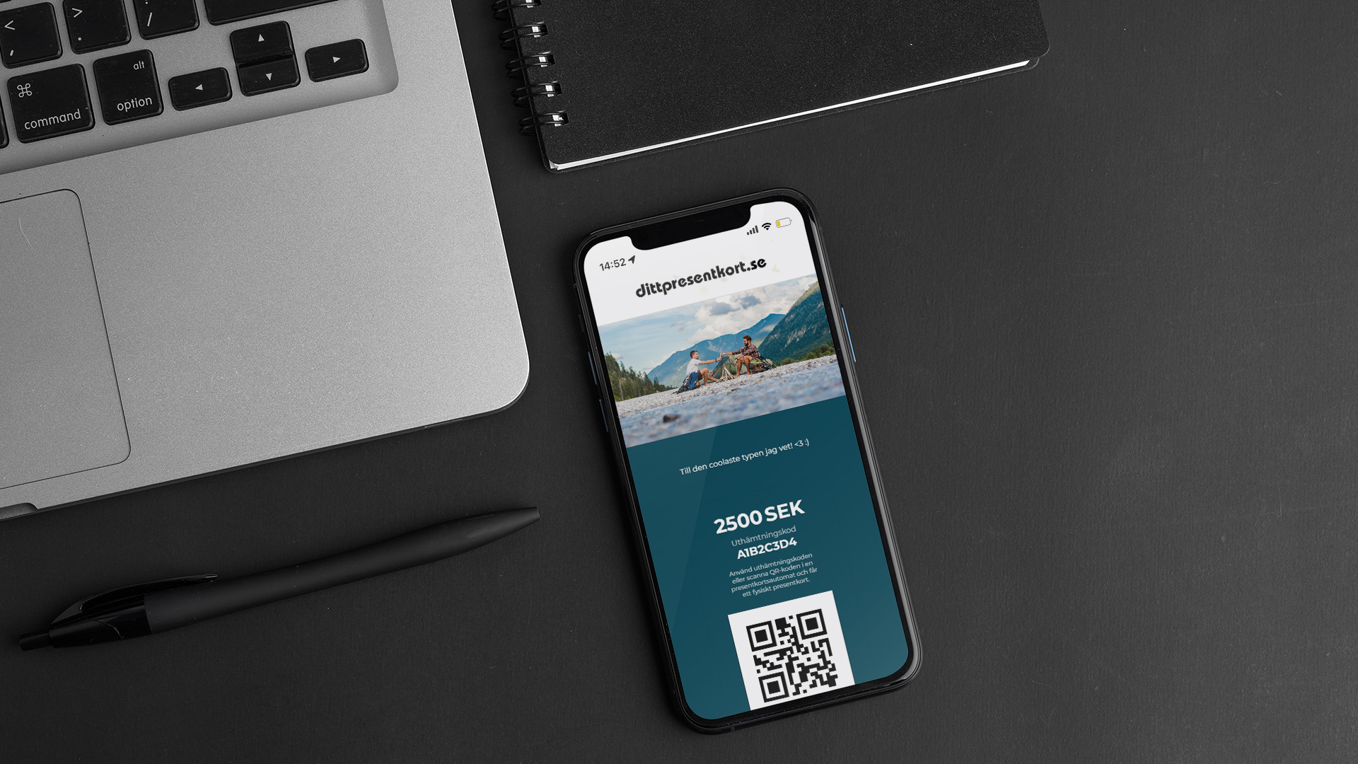 En mobil med dittpresentkort står bredvid en PC på ett svart skrivbord, tillsammans med en svart penna och en svart anteckningsbok.